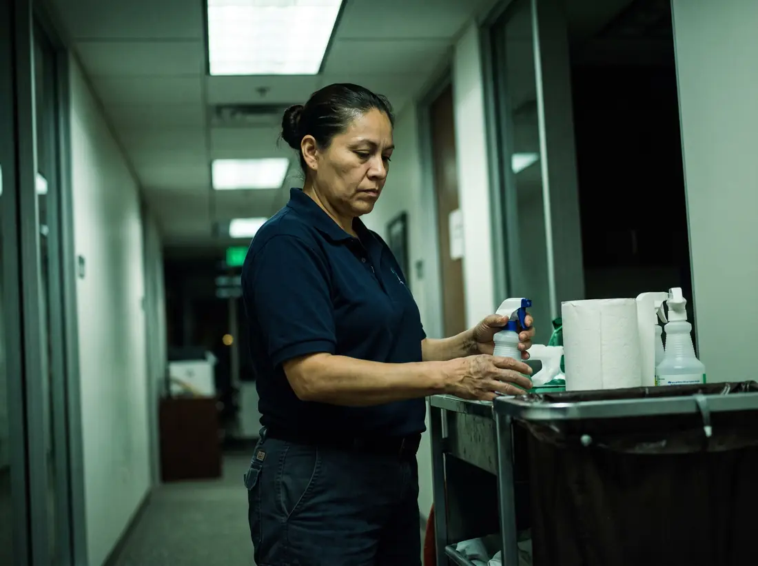 A cleaner restocking a janitorial cart in an office corridor at night.